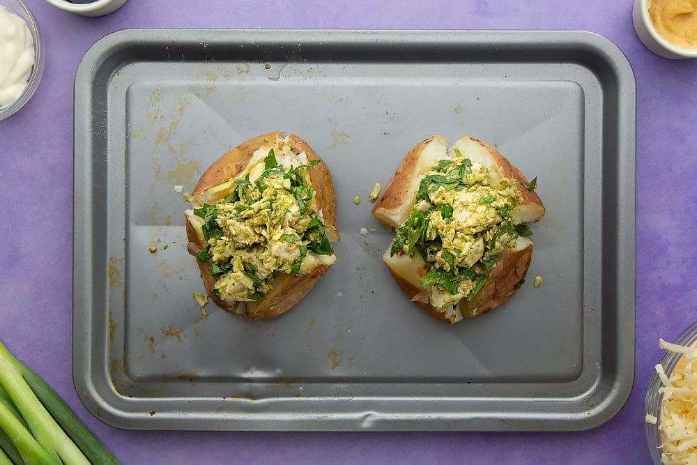 Baked potatoes cooked and cut open on a baking tray, then filled with the cheesy pesto chicken. Ingredients to make jacket potato with cheesy pesto chicken surround the tray.