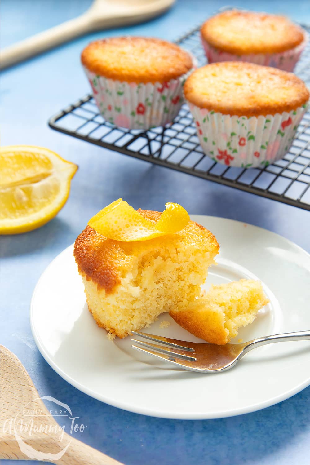 Lemon drizzle cupcake on a white plate with a cake fork. A curl on lemon ring sits on top of the cake, which has been cut open. More cakes are cooling on a rack in the background.