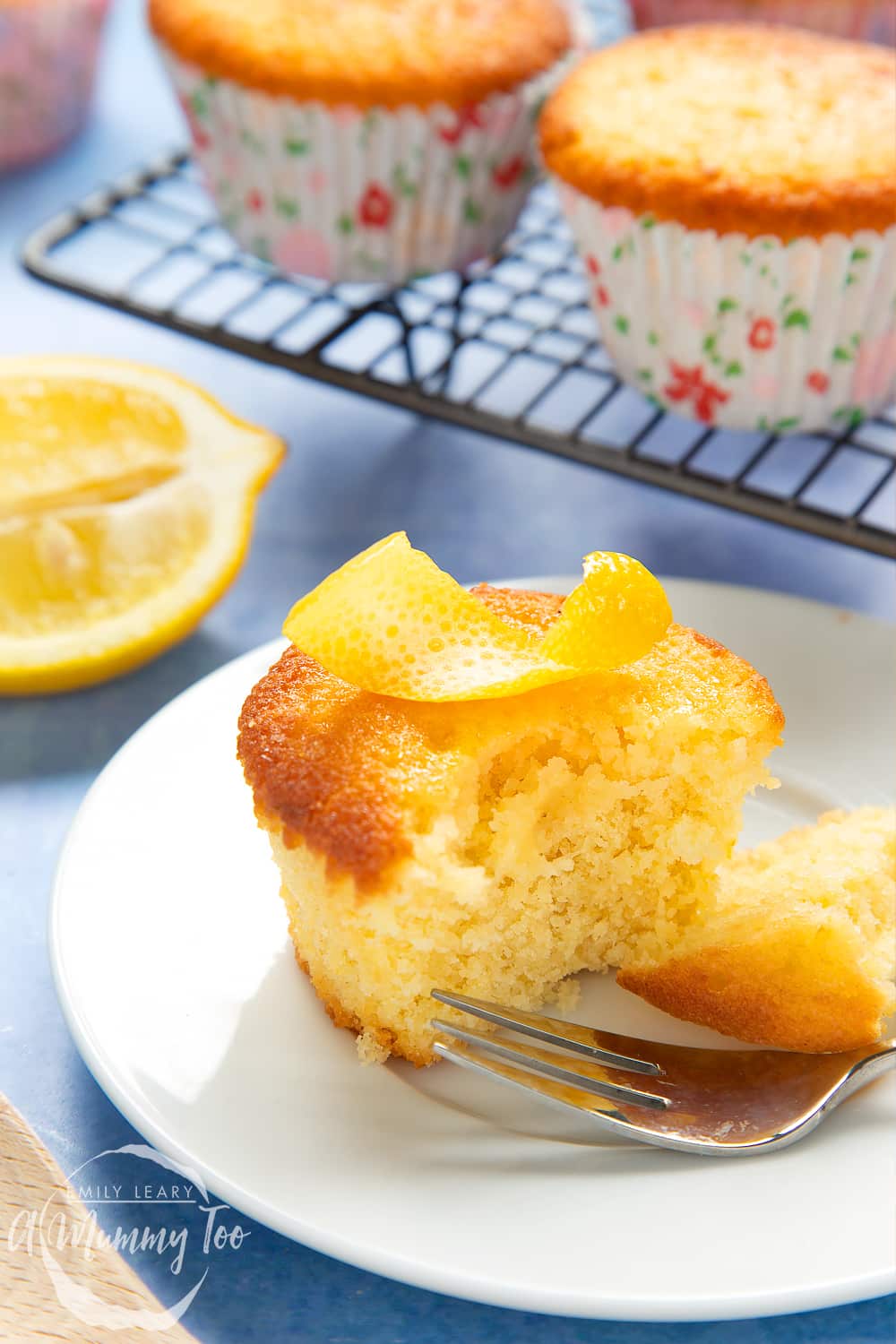 Lemon drizzle cupcake on a white plate with a cake fork. A curl on lemon ring sits on top of the cake, which has been cut open.