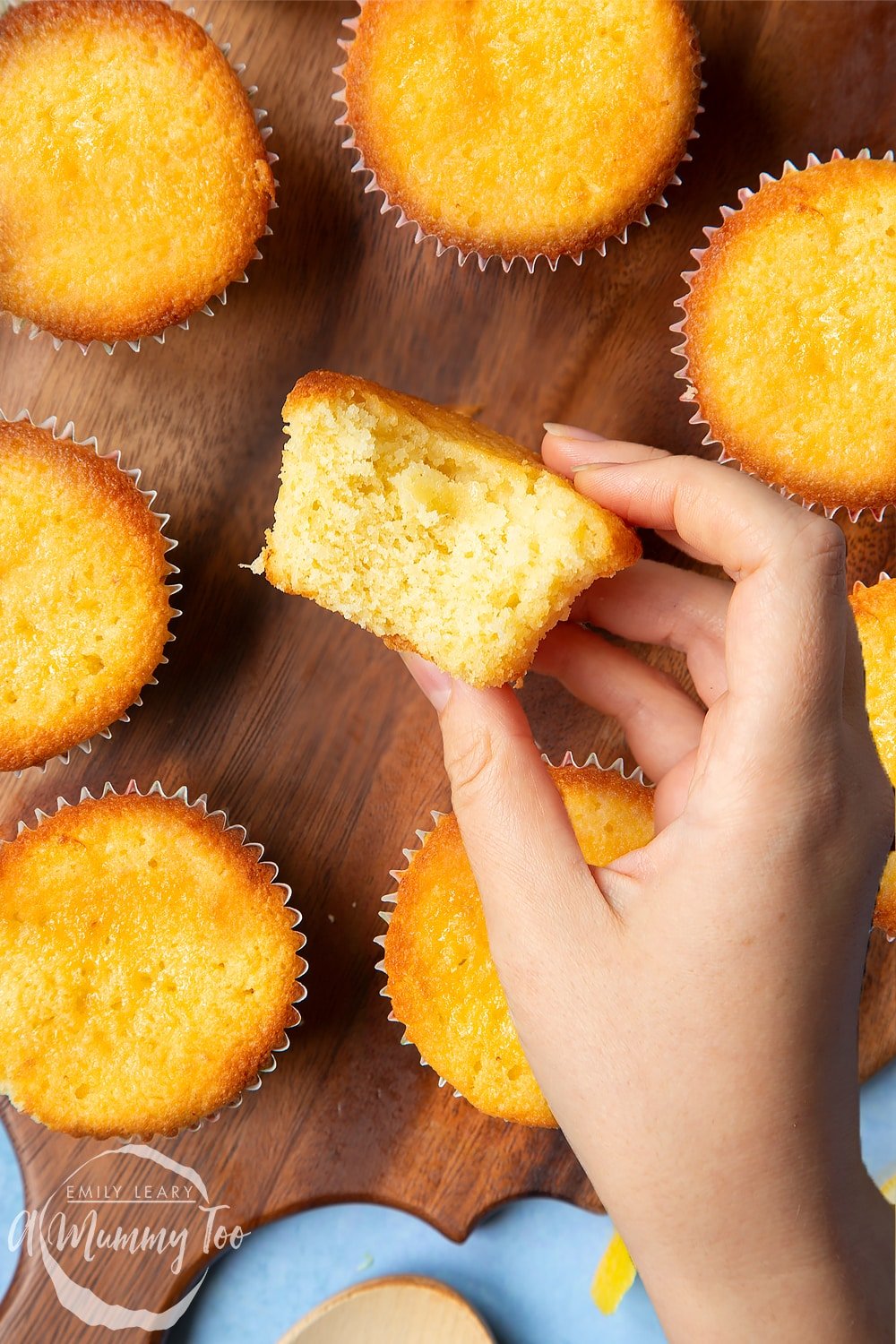 A hand holding a lemon drizzle cupcake with a bite taken out of it. More cupcakes sit on a wooden board in the background.