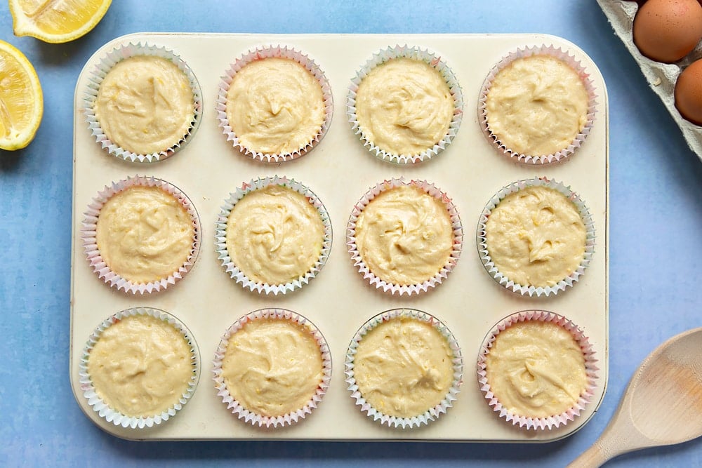 Lemon drizzle cupcake batter in a cream-coloured muffin tray lined with cupcake cases.