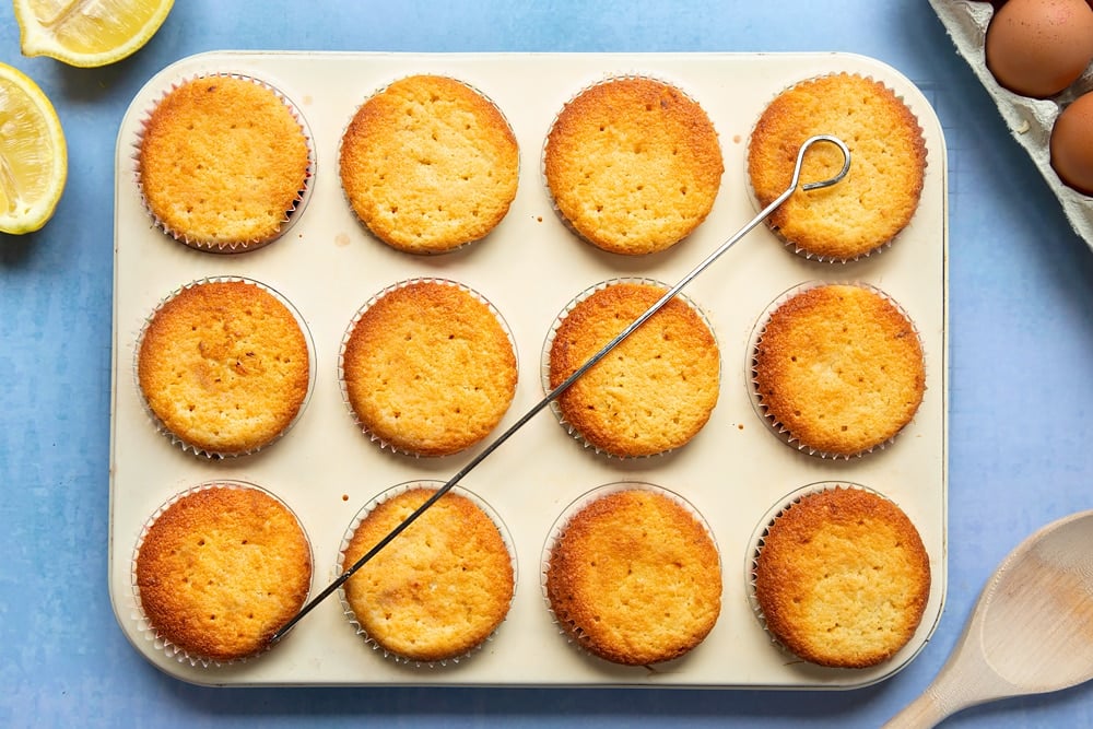 Lemon drizzle cupcakes in a cream-coloured muffin tray. Holes have been poked into the cupcakes with a skewer.