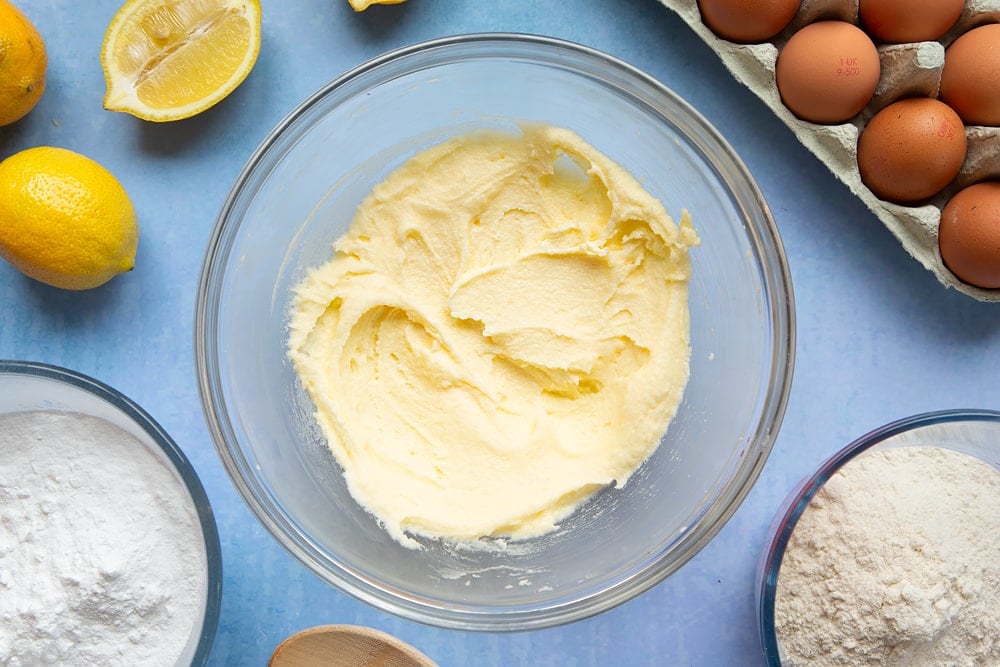 Butter and sugar creamed together in a glass mixing bowl, surrounded by ingredients to make lemon drizzle cupcakes.