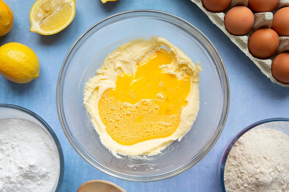 Butter and sugar creamed together in a glass mixing bowl with eggs on top. The bowl is surrounded by ingredients to make lemon drizzle cupcakes.