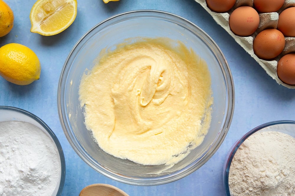 Butter, sugar and eggs creamed together in a glass mixing bowl. The bowl is surrounded by ingredients to make lemon drizzle cupcakes.