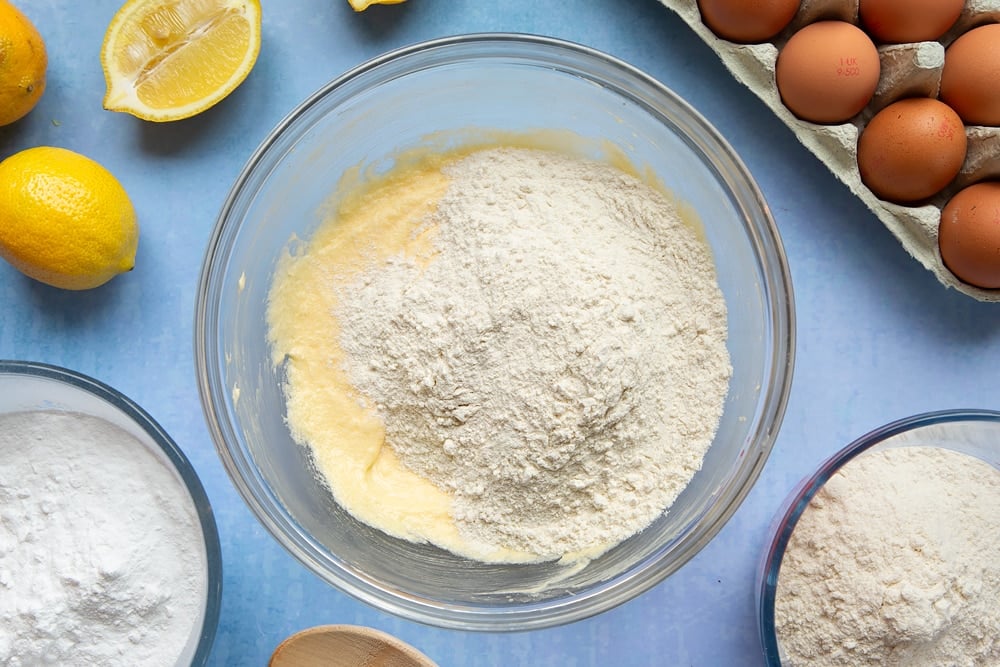 Butter, sugar and eggs creamed together in a glass mixing bowl with flour on top. The bowl is surrounded by ingredients to make lemon drizzle cupcakes.