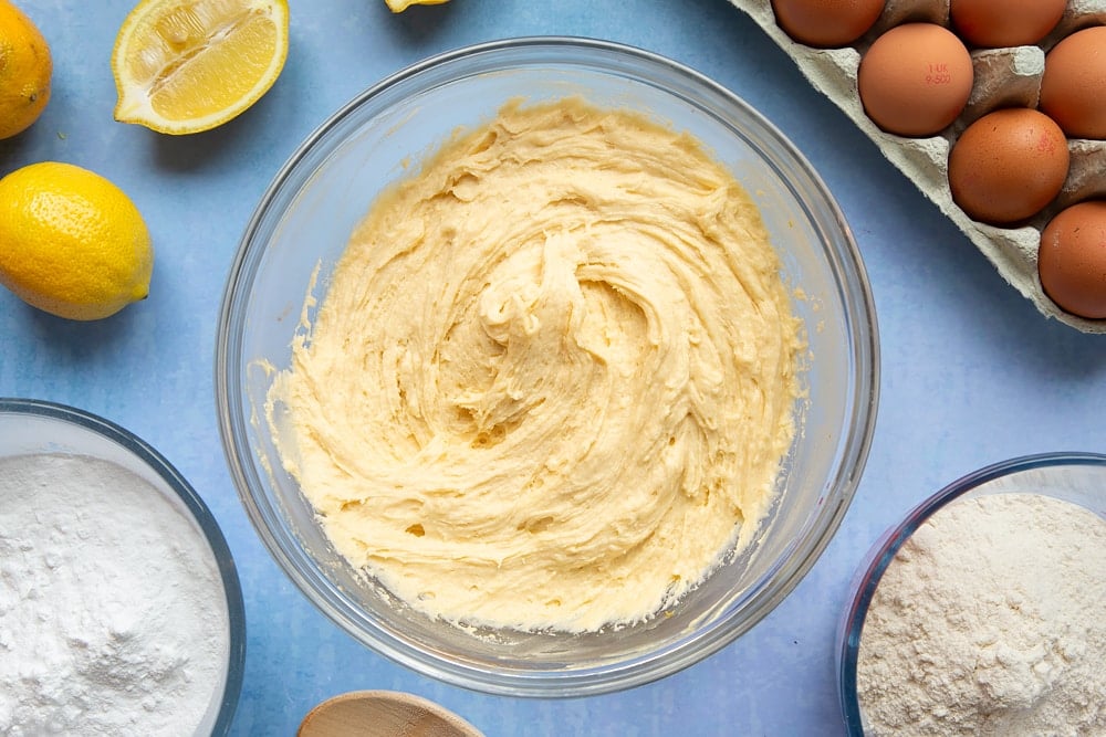 Cake batter in a glass mixing bowl. The bowl is surrounded by ingredients to make lemon drizzle cupcakes.