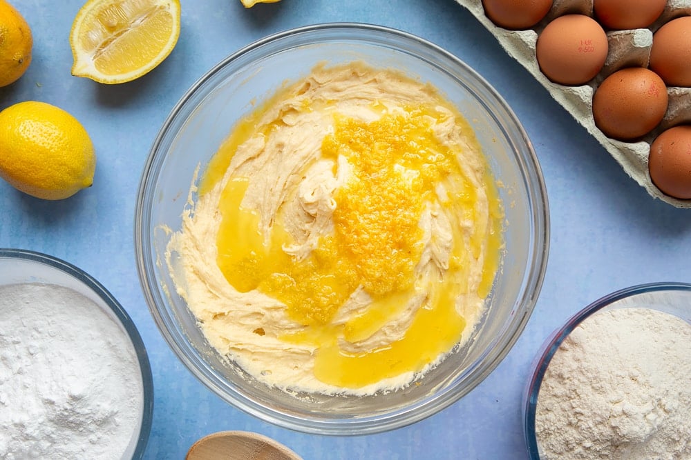 Cake batter in a glass mixing bowl with lemon juice and zest on top. The bowl is surrounded by ingredients to make lemon drizzle cupcakes.