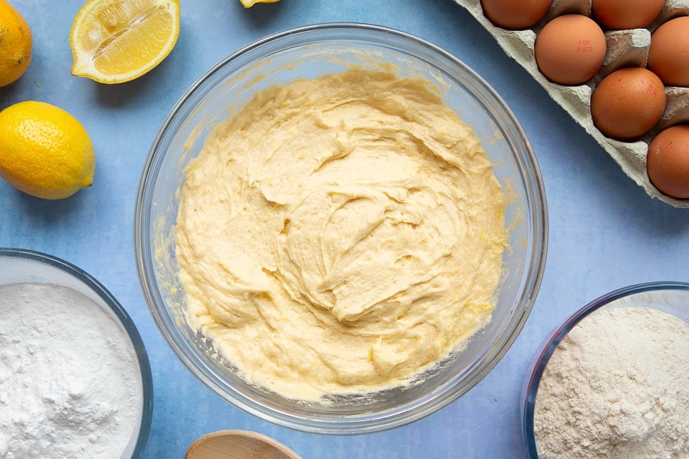 Lemon drizzle cupcake batter in a glass mixing bowl with flour on top. The bowl is surrounded by ingredients to make lemon drizzle cupcakes.