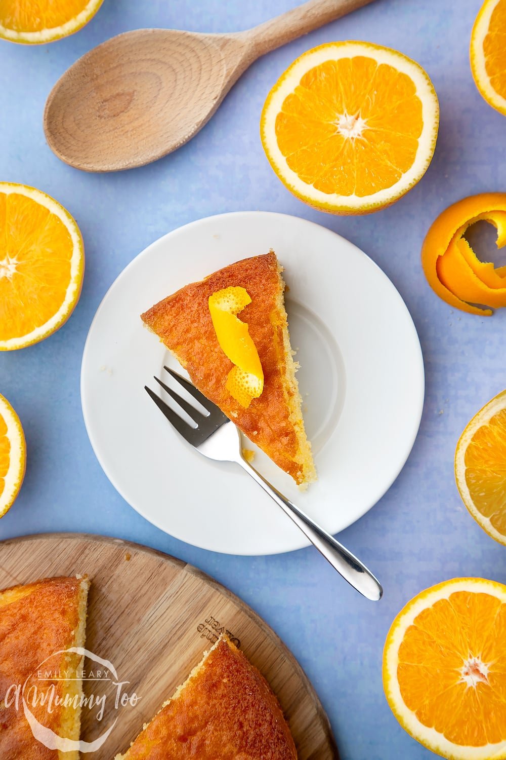 A slice of orange drizzle cake served on a small white plate. A curl of orange peel sits on top of the cake. A cake fork rests on the plate.