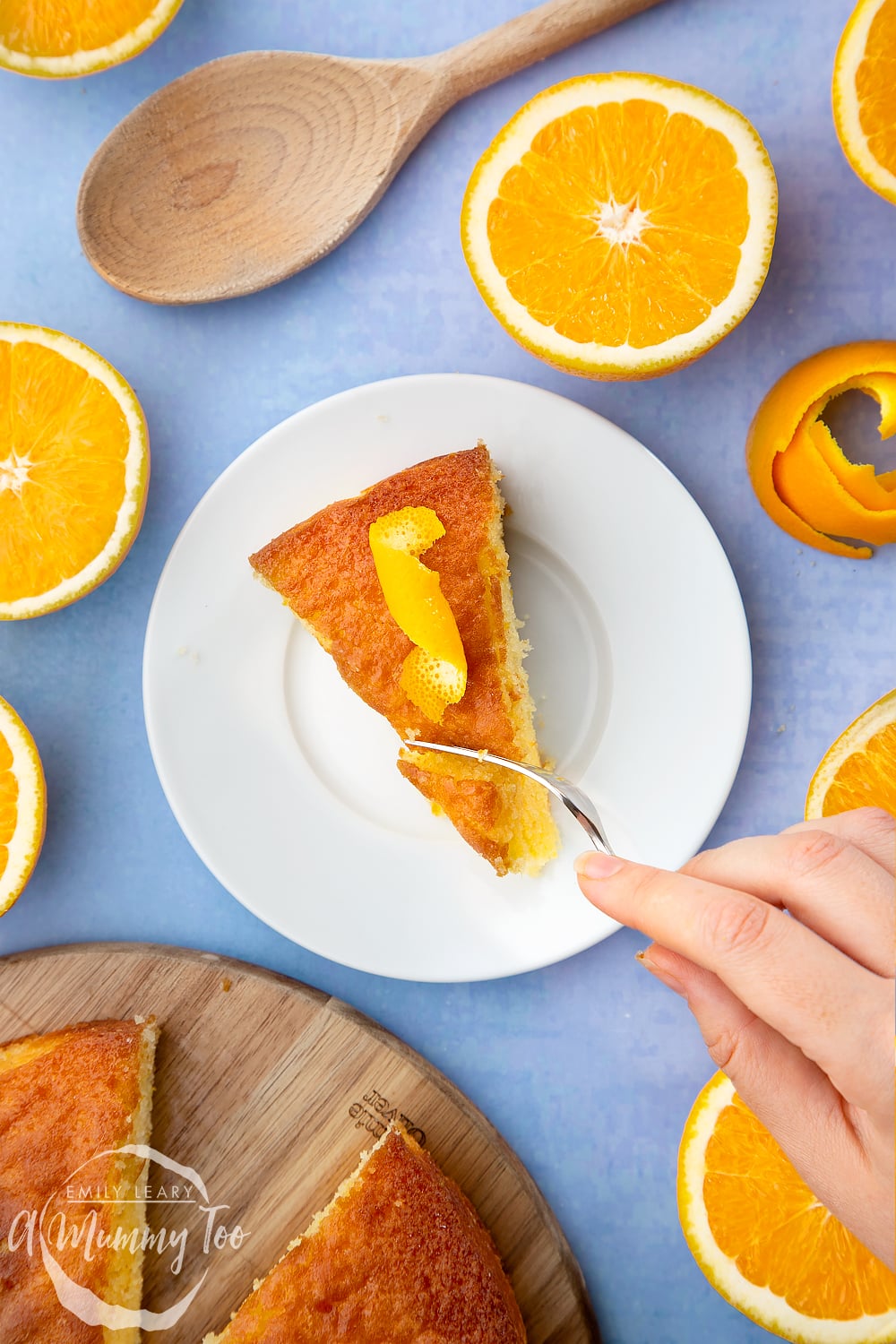 A slice of orange drizzle cake served on a small white plate. A curl of orange peel sits on top of the cake. A hand holding a cake fork cuts into the cake.