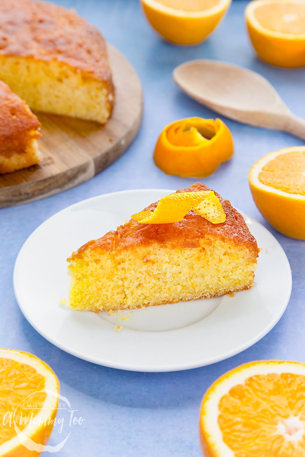 A slice of orange drizzle cake served on a small white plate. A curl of orange peel sits on top of the cake. 