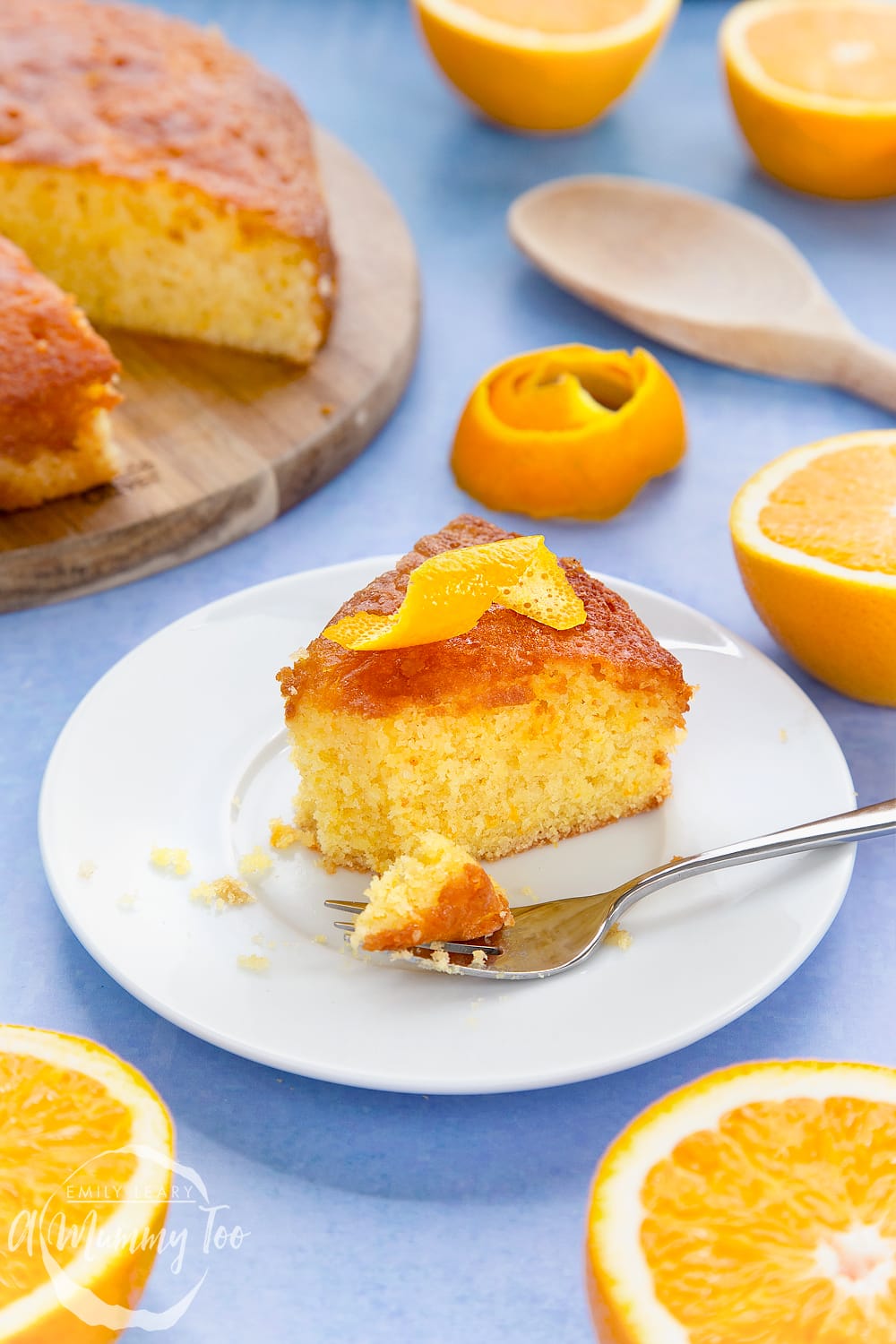 A slice of orange drizzle cake served on a small white plate. A curl of orange peel sits on top of the cake. A cake fork rests on the plate with crumbs on it.