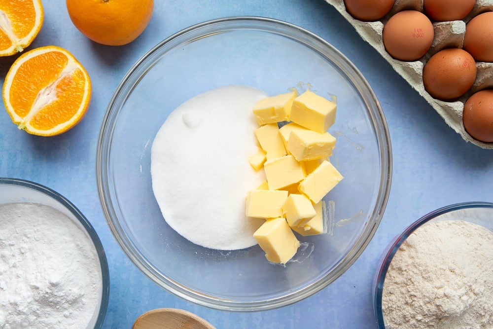 Cubed butter and sugar in a mixing bowl. Ingredients to make orange drizzle cake surround the bowl.