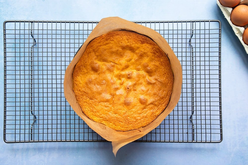 Orange drizzle cake in a greased and lined cake tin, cooling on a wire rack.