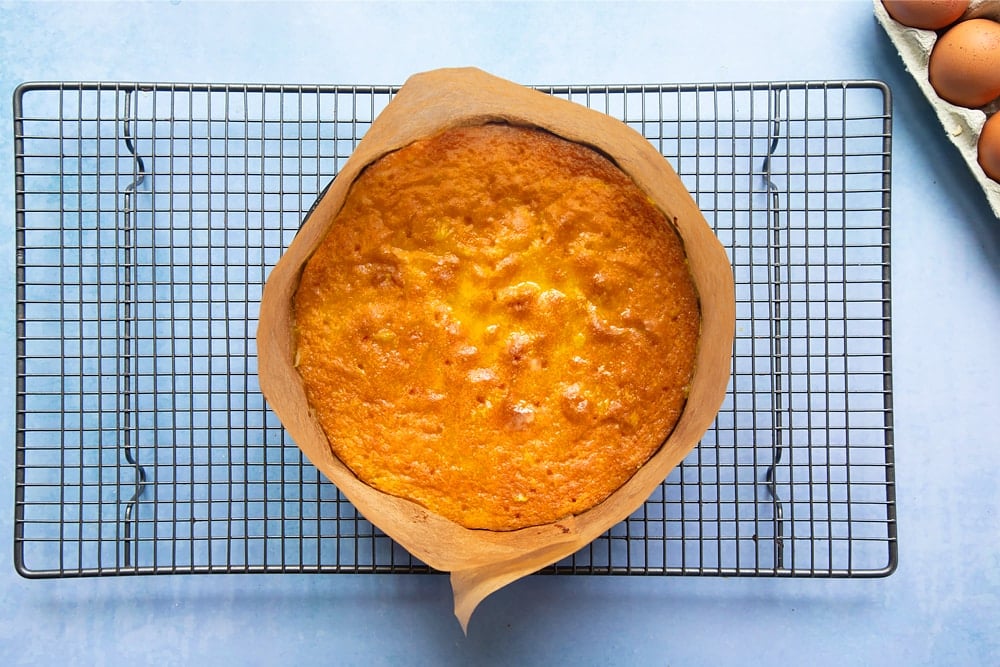 Orange drizzle cake in a greased and lined cake tin, cooling on a wire rack with holes poked into the top and syrup poured on top.