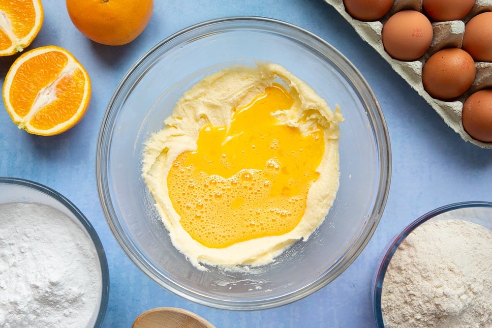 Butter and sugar creamed together in a mixing bowl with beaten egg on top. Ingredients to make orange drizzle cake surround the bowl.