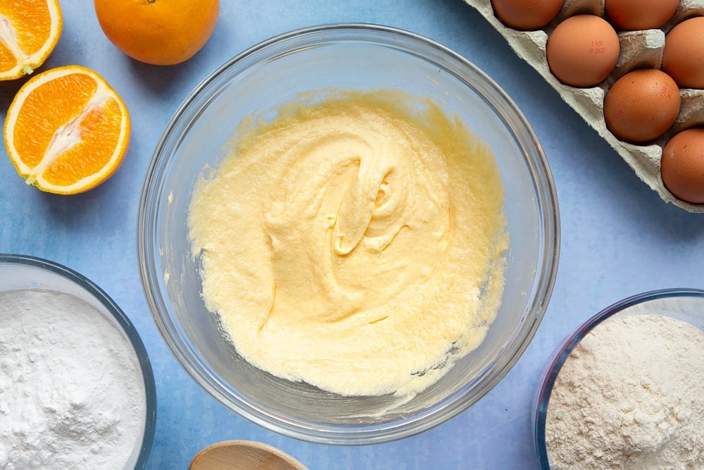 Butter, sugar and eggs creamed together in a mixing bowl. Ingredients to make orange drizzle cake surround the bowl.