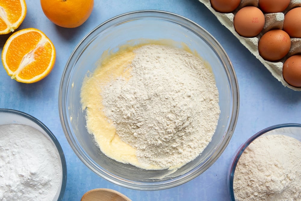 Butter, sugar and eggs creamed together in a mixing bowl with self-raising flour on top. Ingredients to make orange drizzle cake surround the bowl.
