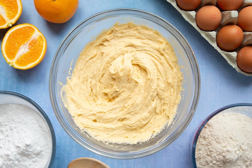 Cake batter in a mixing bowl. Ingredients to make orange drizzle cake surround the bowl.