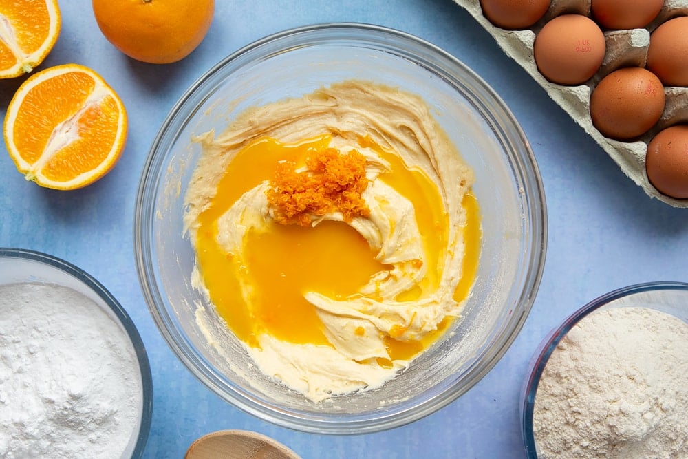 Cake batter in a mixing bowl with orange juice and zest on top. Ingredients to make orange drizzle cake surround the bowl.