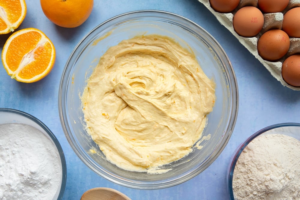 Orange drizzle cake batter in a mixing bowl. Ingredients to make orange drizzle cake surround the bowl.