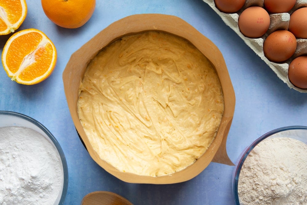 Orange drizzle cake batter in a greased and lined cake tin. Ingredients to make orange drizzle cake surround the tin.