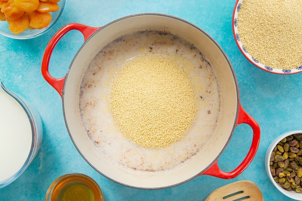 Overhead shot of adding the cous cous to the pan of ingredients. The pan sits on a blue background surrounded by additional ingredients required for the recipe. 