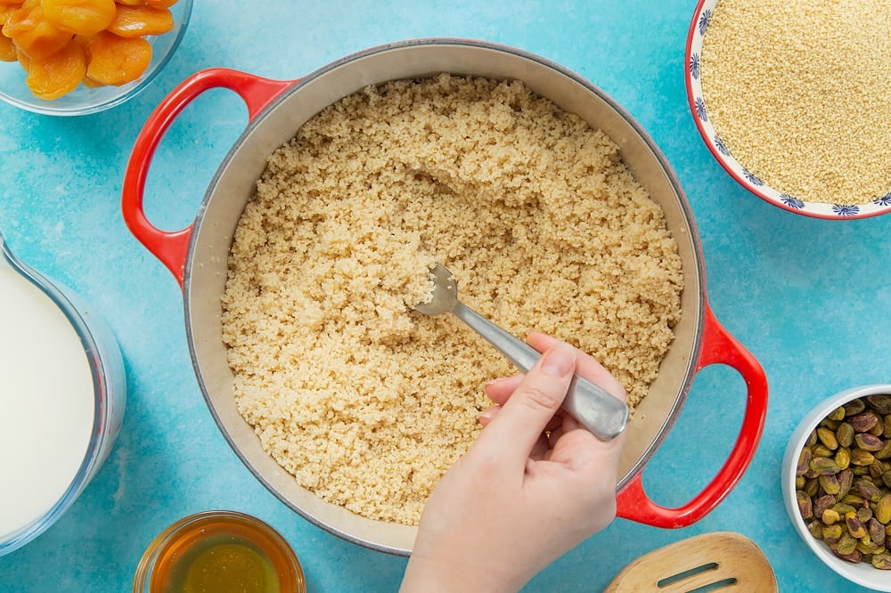 Fluffing the sweet breakfast couscous after it's been steamed with the lid on. The fluffing is being done with a fork. At the side of the pan there's additional ingredients used for this recipe. 