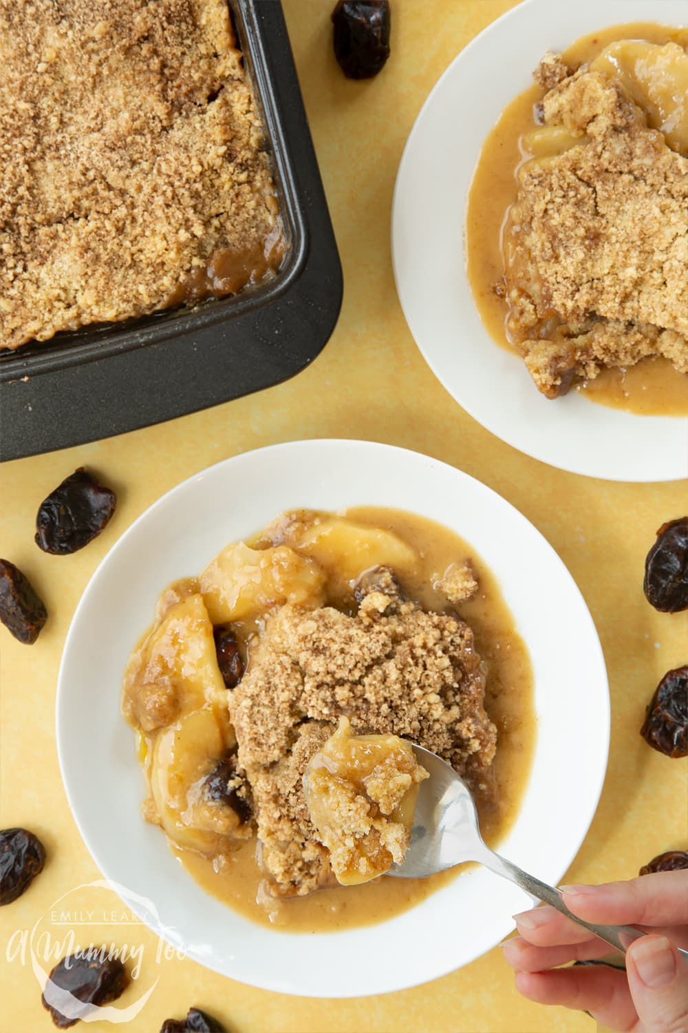 Overhead shot of a spoon going into a plate of toffee apple crumble. At the side there's an additional plate of toffee apple crumble and a pan.