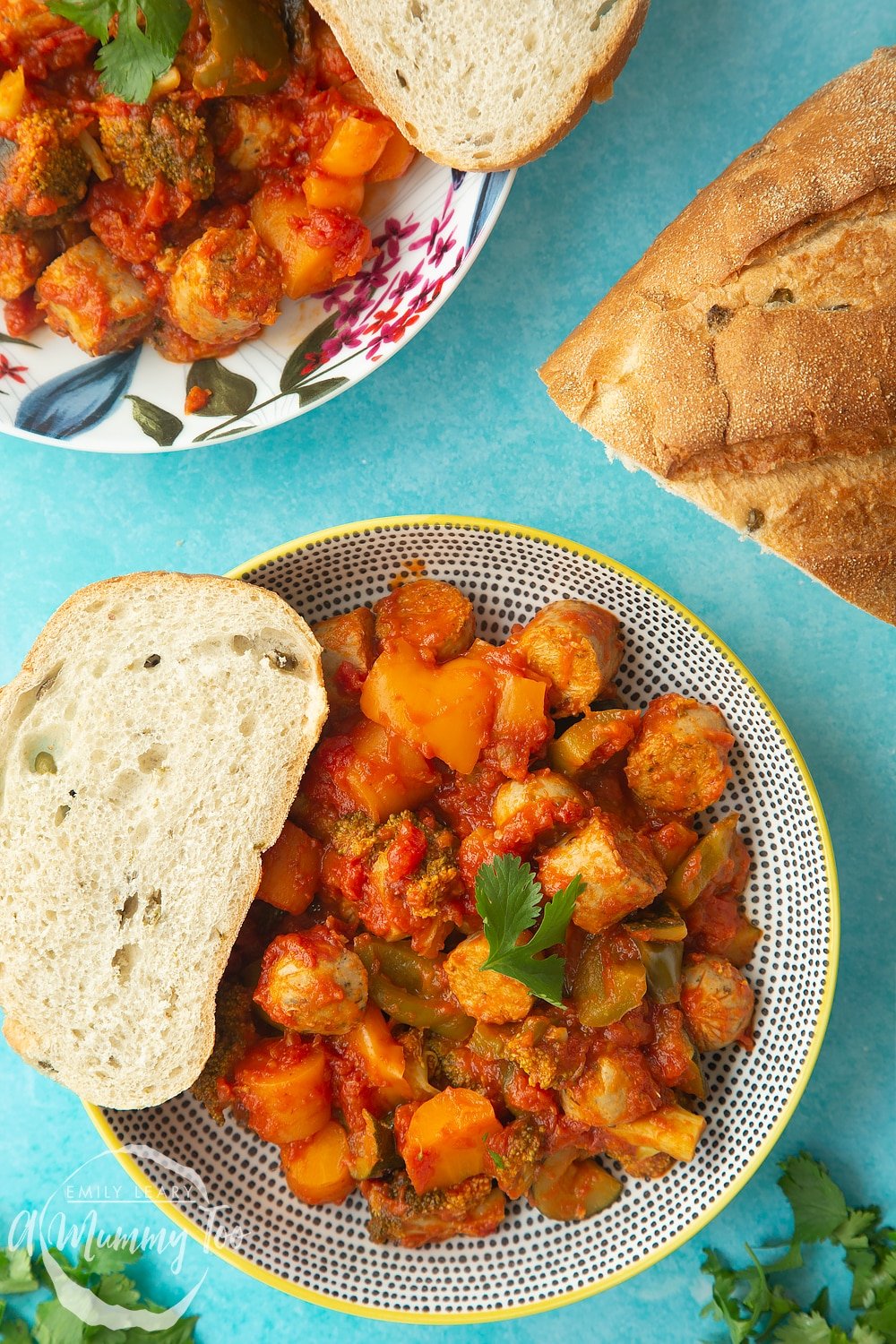 Two bowls of slow-cooked vegetarian sausage casserole on a blue background with a crusty bread roll at the side. 