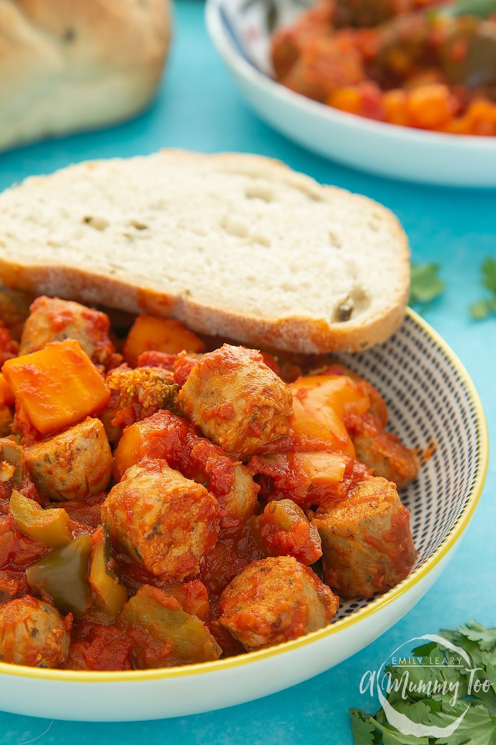 Close up of a bowl of slow-cooked vegetarian sausage casserole with a crusty bread roll on the side. The bowl sits on a blue table. 