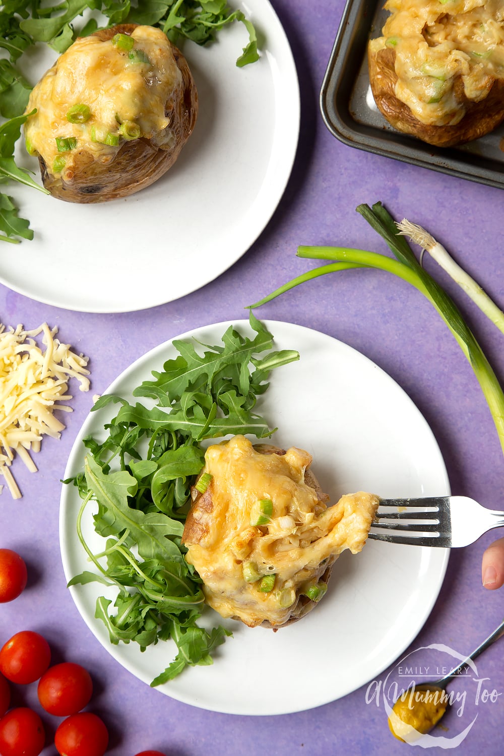 A cheese and onion jacket potato served on a white plate with rocket. A fork stretches the cheese from the potato. Another plate is shown to the side.