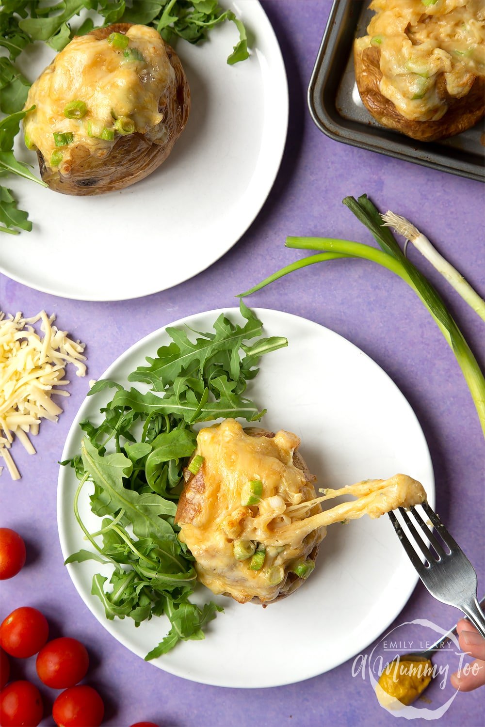 A cheese and onion jacket potato served on a white plate with rocket. A fork stretches the cheese from the potato. More ingredients are shown round the edges.