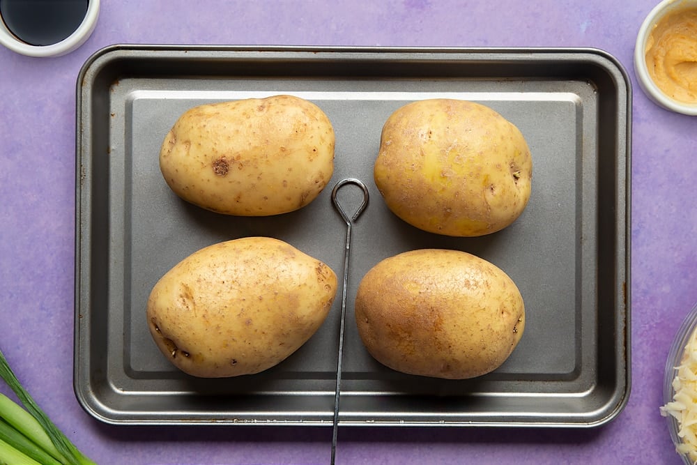 Uncooked jacket potatoes on a tray with a skewer. Ingredients to make cheese and onion jacket potatoes surround the tray.