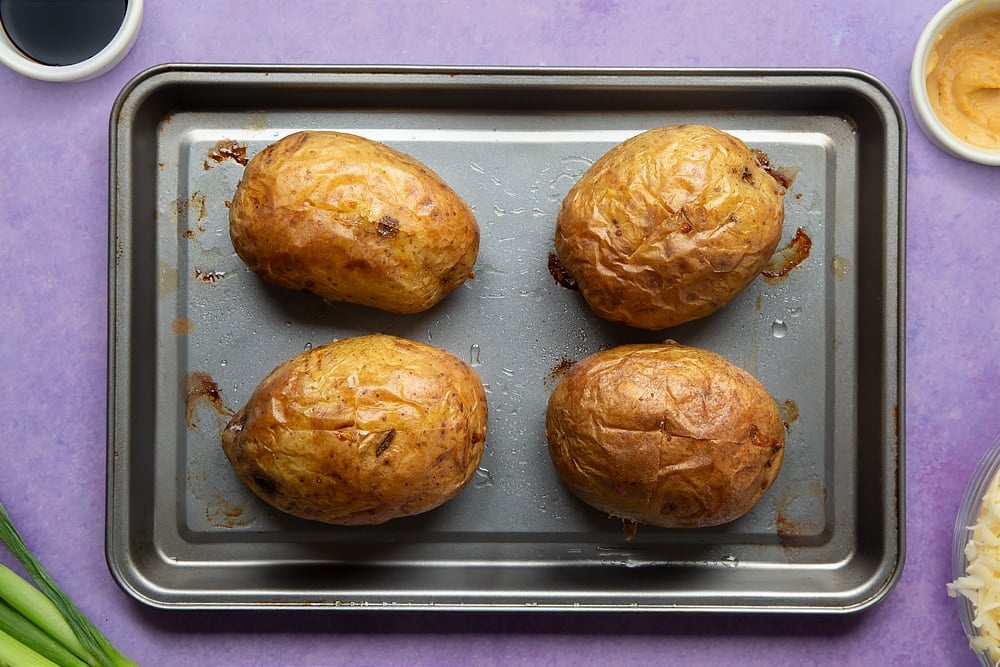 Baked jacket potatoes on a tray. Ingredients to make cheese and onion jacket potatoes surround the tray.