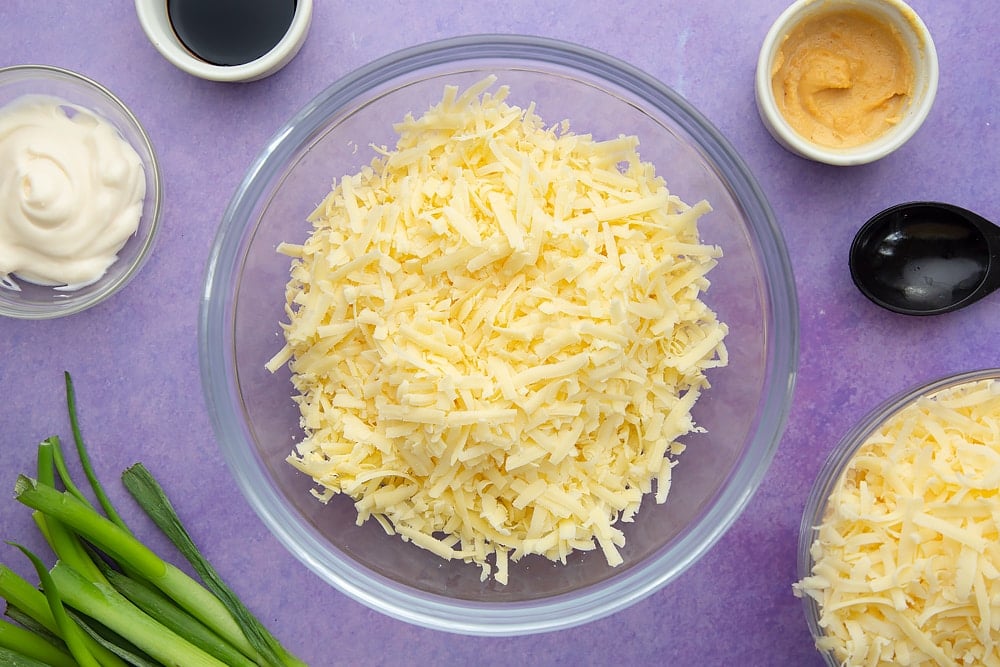 Grated mozzarella and cheddar in a mixing bowl. Ingredients to make cheese and onion jacket potatoes surround the bowl.