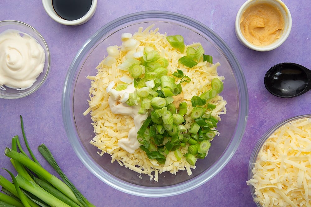 Grated mozzarella, cheddar, spring onions, mayonnaise, mustard and soy sauce in a mixing bowl. Ingredients to make cheese and onion jacket potatoes surround the bowl.