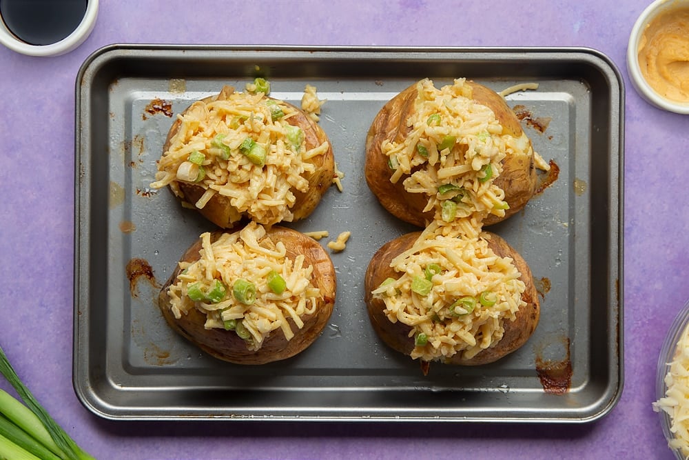 Baked jacket potatoes on a tray topped with a cheese and onion mix. Ingredients to make cheese and onion jacket potatoes surround the tray.