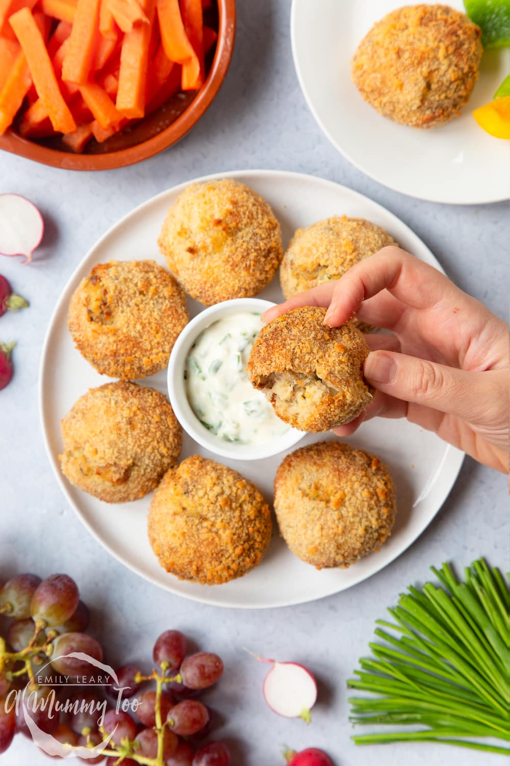 A hand holding a Omega 3 rich smoked mackerel fishcake on a white plate with a chive dip in the center.