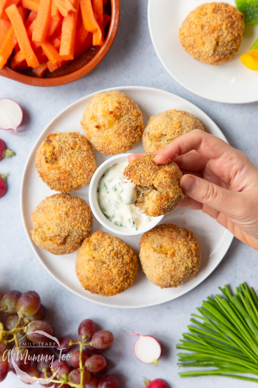 An omega 3 rich smoked mackerel fishcake being held by a hand over a plate of additional fishcakes. At the side there's some ingredients that are used to make the fishcakes.