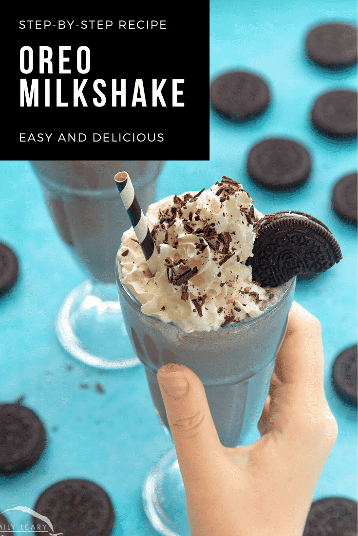 A hand holding an oreo milkshake topped with whipped cream and oreo cookies with a black and white striped paper straw. In the background there's some additional oreo cookies and another oreo milkshake. In the top right corner there's some text describing the image for Pinterest. 