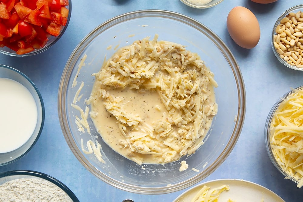 Mixing together some of the cheese and red pepper muffins ingredients. The bowl sits on a light blue background with additional ingredients scatted around the side.