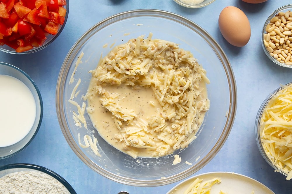 Mixing together some of the cheese and red pepper muffins ingredients. The bowl sits on a light blue background with additional ingredients scatted around the side. 