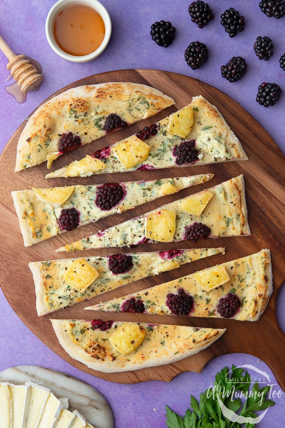 Brie and blackberry pizza sliced on a wooden board. Ingredients surround the board.