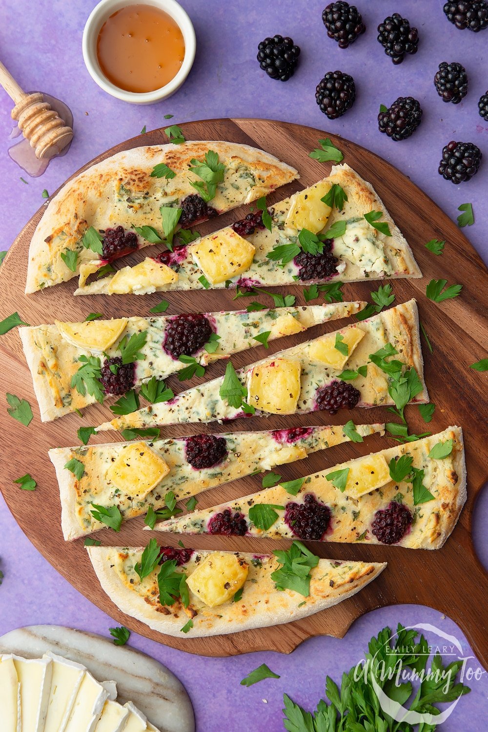 Brie and blackberry pizza sliced on a wooden board and scattered with fresh parsley. Ingredients surround the board.