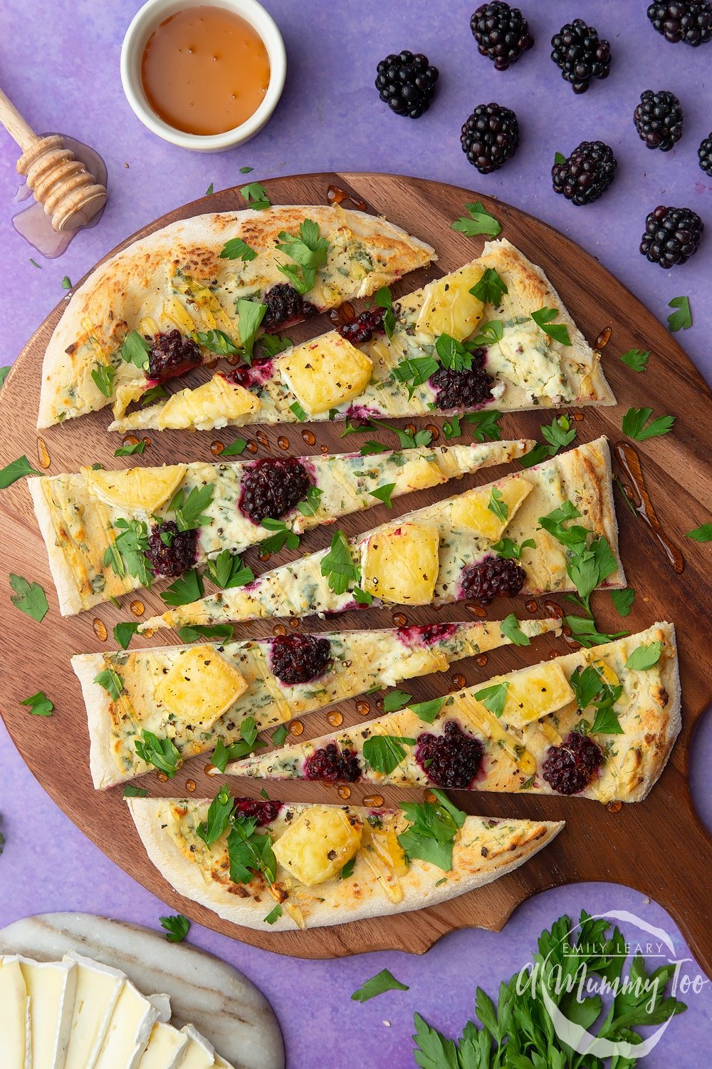 Brie and blackberry pizza sliced on a wooden board and scattered with fresh parsley and drizzled with honey. Ingredients surround the board.