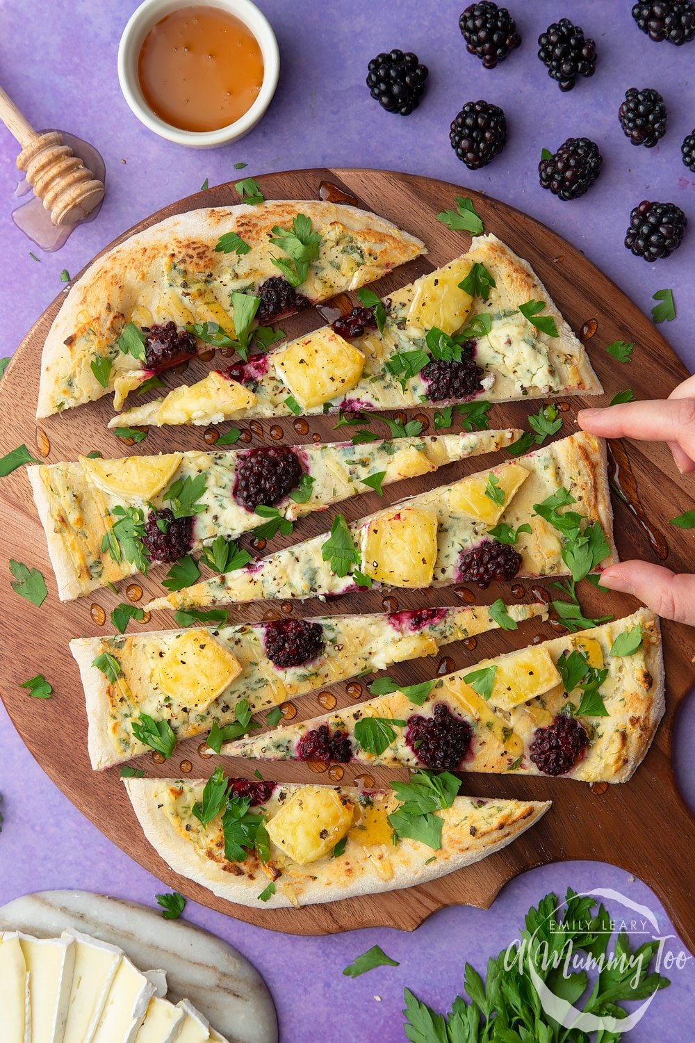 Brie and blackberry pizza sliced on a dark wooden board. A hand reaches in to take a slice.