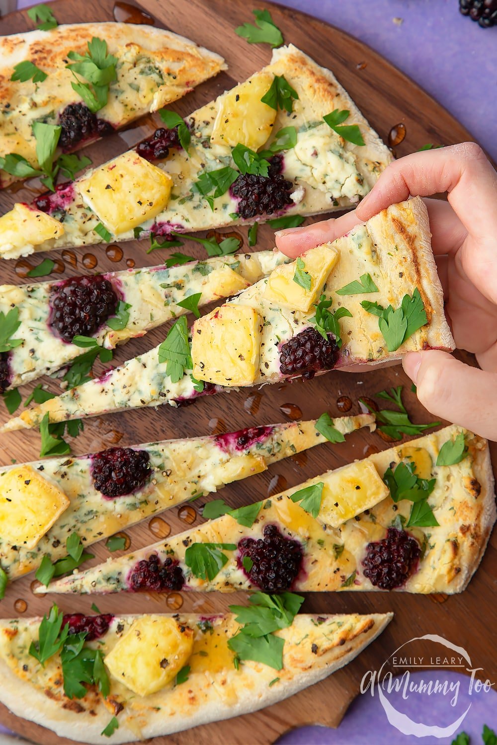 A hand picking up a slice of brie and blackberry pizza on a dark wooden board.