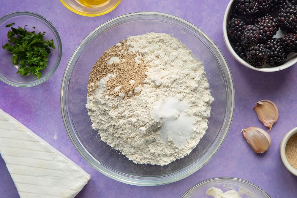 Flour, yeast and salt in a glass mixing bowl. Ingredients for brie and blackberry pizza surround the bowl.
